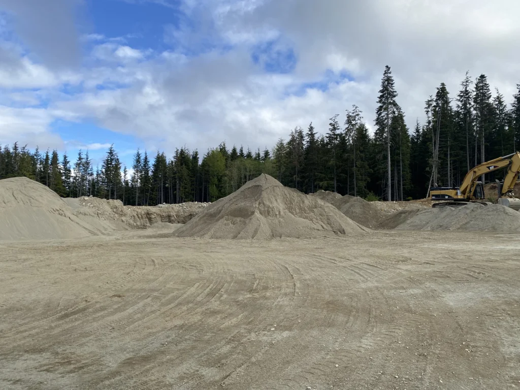 Multiple gravel and sand stockpiles at an aggregate pit, with an excavator and forest in the background.